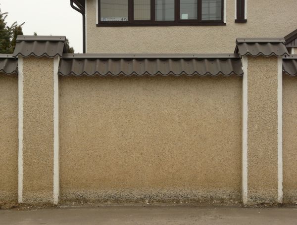 Concrete fence texture made up of a solid tan wall with a coarse, fine texture, similar columns with white trim on either side, and a dark brown tile awning along the top. Discoloration is visible near the bottom of the wall.